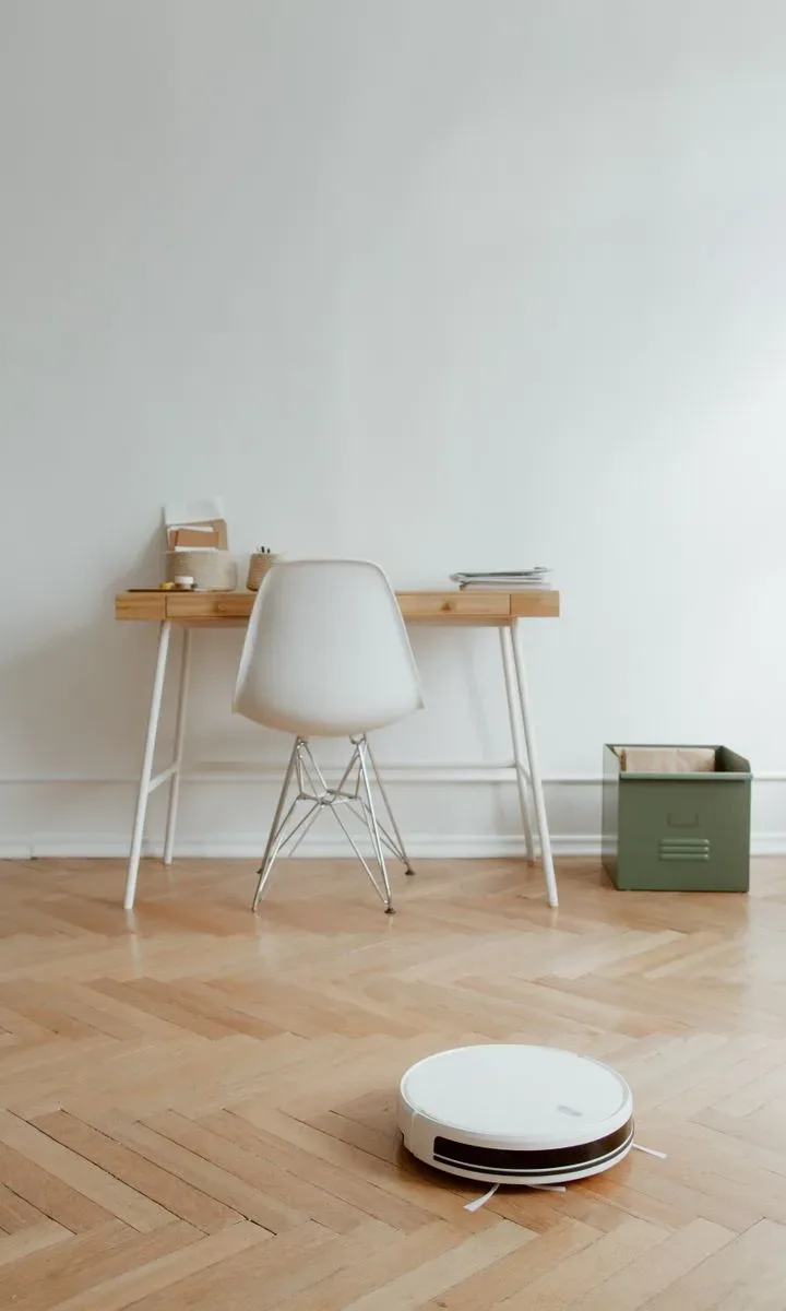 10 productos de limpieza económicos que siempre deberías tener en casa. Aspirador robot automático limpiando el suelo de una sala de estar, destacando un producto básico para la limpieza del hogar.