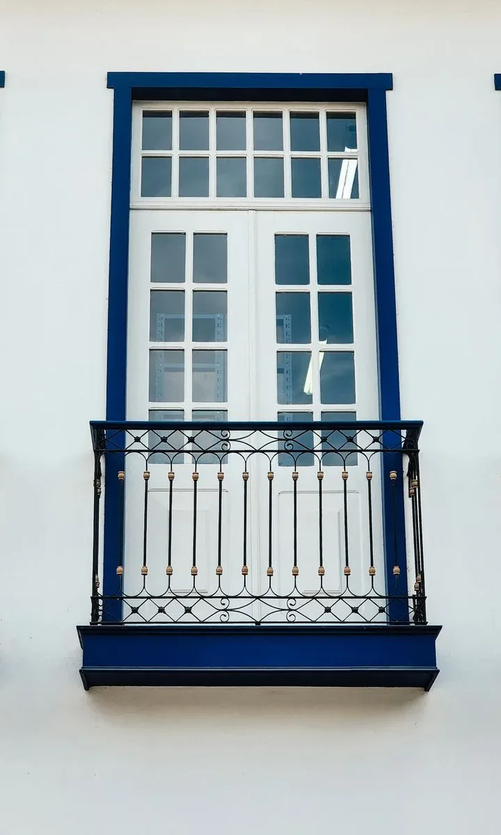 Persona limpiando una barandilla de vidrio en un balcón con un paño y spray, con vista a la ciudad, representando la limpieza de ventanas y barandillas.