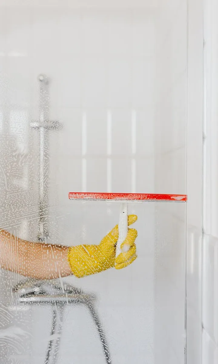 Cómo quitar olor a humedad en casa en pocos paso: 6 trucos naturales y caseros Una persona limpiando la mampara de la ducha con papel de horno mojado para eliminar la cal y prevenir el olor a humedad en el baño.